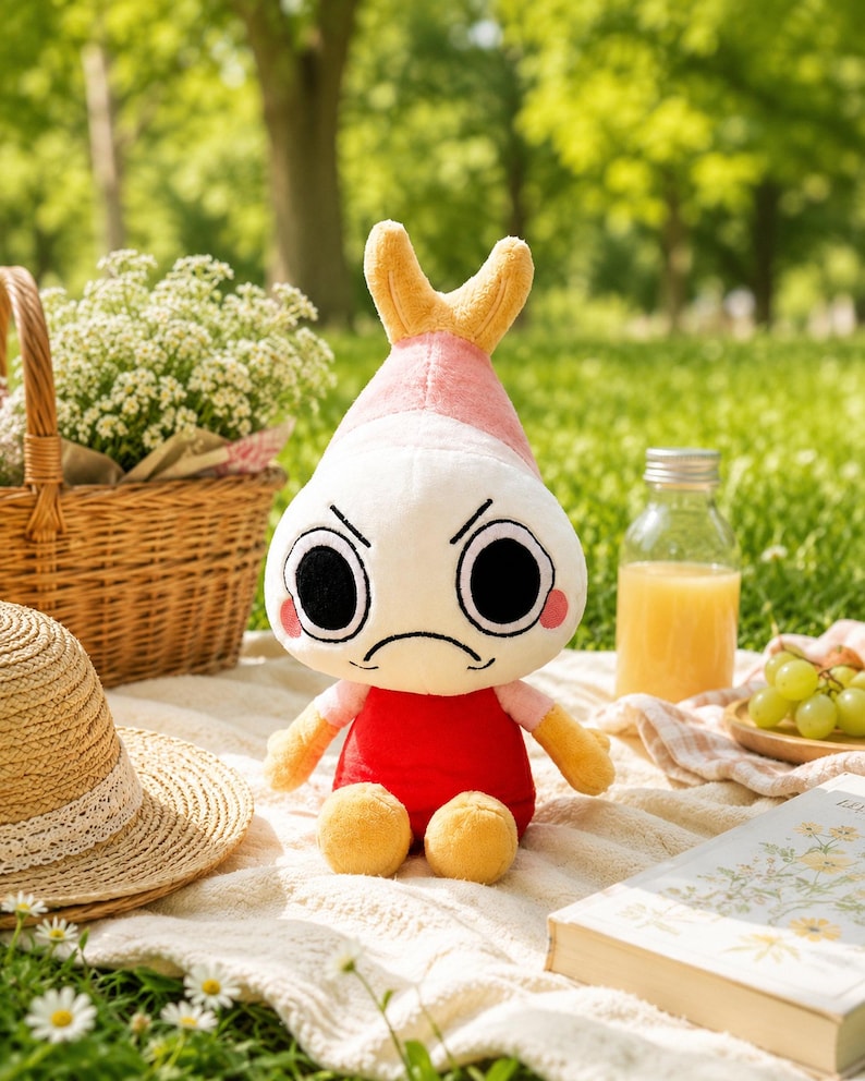 Puede incluir: Un peluche con cuerpo blanco, camisa roja y pies y brazos amarillos. El juguete tiene una cabeza rosa y amarilla con una expresi&oacute;n de mal humor y grandes ojos negros. El juguete est&aacute; sentado en una manta blanca con una cesta de picnic, un sombrero y un libro en el fondo.