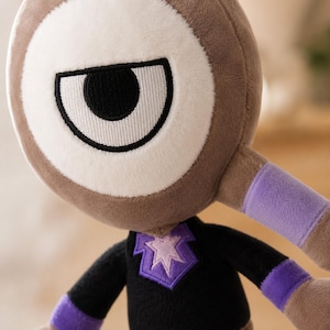 May include: A plush toy with a large, stylized eye. The toy has a brown body, a white and brown eye, and a black shirt with a purple star design. Purple bands are on the arms. The toy is soft and cuddly.