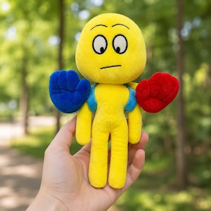 Puede incluir: Un peluche amarillo con cabeza redonda, ojos grandes y expresión neutra. Tiene accesorios en forma de mano azules y rojos. El juguete se sostiene en una mano sobre un fondo verde borroso.