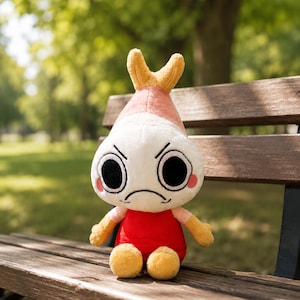 Puede incluir: Un peluche de camar&oacute;n con cabeza blanca, cuerpo rojo y detalles amarillos. El camar&oacute;n tiene una expresi&oacute;n de enfado con grandes ojos negros y mejillas rosadas. Est&aacute; sentado en un banco de madera en un parque.