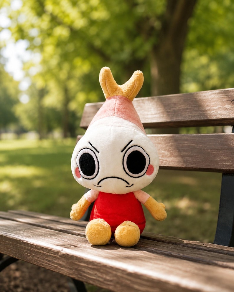 Puede incluir: Un peluche de camar&oacute;n con cabeza blanca, cuerpo rojo y detalles amarillos. El camar&oacute;n tiene una expresi&oacute;n de enfado con grandes ojos negros y mejillas rosadas. Est&aacute; sentado en un banco de madera en un parque.