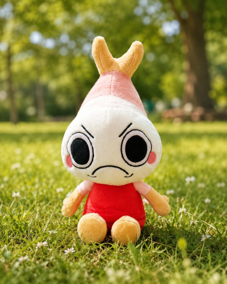 Puede incluir: Un peluche con forma de camar&oacute;n con una expresi&oacute;n de enfado. El juguete tiene una cabeza blanca, una parte superior rosa y amarilla, un cuerpo rojo y brazos y piernas peque&ntilde;os. Est&aacute; sobre hierba verde.