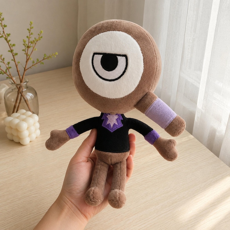 May include: A plush toy with a large, brown, circular head featuring a white eye with a black pupil. The toy has a black shirt with a purple collar and a purple band on its arm. The toy is brown and has small arms and legs.