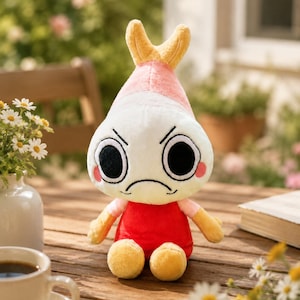 Puede incluir: Un peluche con forma de camar&oacute;n con una expresi&oacute;n de mal humor. El juguete tiene una cabeza blanca con ojos negros, una parte superior rosa y amarilla, un cuerpo rojo y brazos y pies amarillos. Est&aacute; sentado en una mesa de madera.