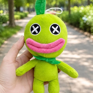 May include: A bright green plush toy with a round head, a single leaf on top, and a white hanging loop. The face features two black and white stitched X-shaped eyes and a wide, textured pink mouth. A small green fabric collar sits at the neck.