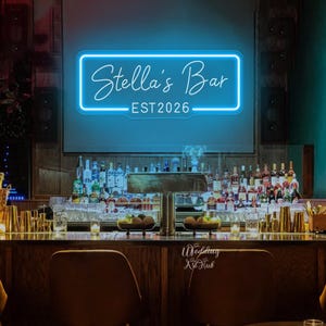 Puede incluir: Un letrero de neón rectangular con un brillo azul intenso montado en una pared. El letrero presenta el texto Stella's Bar en una fuente cursiva, con el texto EST 2026 impreso en una fuente sans-serif debajo, todo dentro de un borde de neón azul.