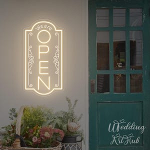 May include: A glowing neon sign with the words "We are OPEN" in a decorative frame. The sign is mounted on a white wall next to a green door. A basket of flowers and plants sits below the sign.