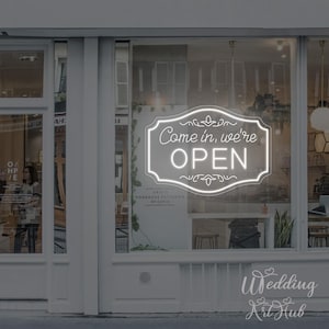 Op de afbeelding: Een wit neonbord in een etalage leest "Come in, we're OPEN". Het bord staat in een decoratief kader. De etalage toont ook de tekst "OBJETS HOMEMADE PATISSERIE SPICERIE."