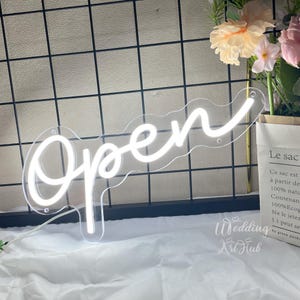 Pode incluir: Um letreiro de néon branco com a palavra Open numa fonte cursiva. O letreiro está montado sobre uma base de acrílico transparente com orifícios de montagem visíveis. A luz emite um brilho intenso em frente a um painel de grelha preto.