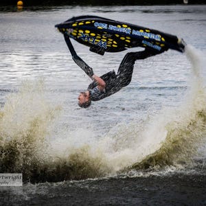 May include: A jet ski performs a mid-air flip, creating a large splash of water. The jet ski is black and yellow, and the rider is wearing a dark wetsuit. The water is choppy, and the sky is overcast.