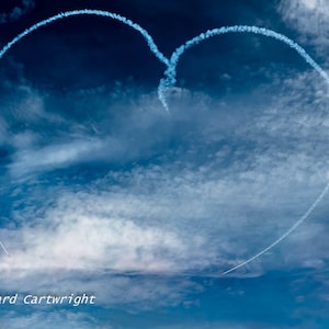 May include: A heart shape formed by a contrail in a blue sky with scattered clouds. The image includes the text "Richard Cartwright" in the bottom left corner. The sky is a gradient of blues.