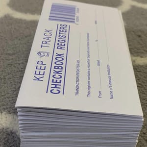 May include: A stack of white checkbook registers with blue text that reads "KEEP TRACK CHECKBOOK REGISTERS." The registers include spaces for transaction details and are designed to record deposits and items numbered.