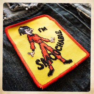 May include: A yellow and red embroidered patch with a cartoon figure in a red jumpsuit and the text "I'm Shootable".