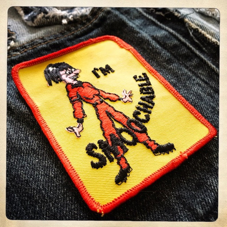 May include: A yellow and red embroidered patch with a cartoon figure in a red jumpsuit and the text "I'm Shootable".