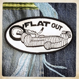 May include: A white oval patch with black stitching and the text "FLAT OUT" in black. A black and white illustration of a motorcycle with a rider is on the patch.