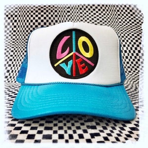Puede incluir: Una gorra de camionero azul y blanca con un símbolo de paz negro bordado en la parte delantera. El símbolo de paz tiene la palabra "LOVE" escrita en su interior con letras de colores.