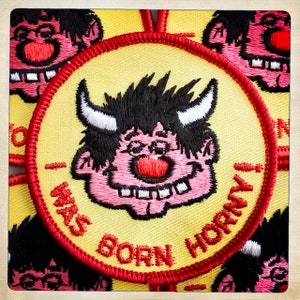 May include: A yellow and red embroidered patch with a cartoon character with black hair, pink skin, and a red nose. The character has a wide smile and white teeth. The text on the patch reads "I Was Born Horny".