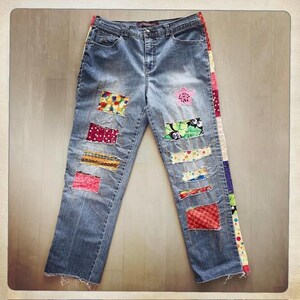 May include: Blue denim jeans featuring a variety of colorful fabric patches sewn onto the front legs. The outer side seam is accented with a vertical strip of multi-colored patchwork fabric. The jeans have a classic five-pocket design.