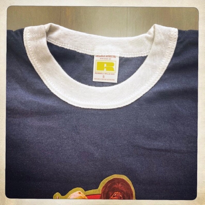 Op de afbeelding: Een donkerblauw T-shirt met een witte boord. Het shirt heeft een label met de tekst "Russell Athletic" en "Russell Mills Inc.". Het label heeft ook een maat "S" erop gedrukt.