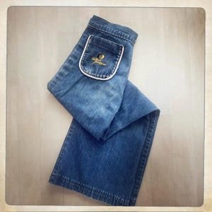 Könnte beinhalten: Eine mittelblaue Jeans aus Denim mit einer aufgesetzten Tasche auf der Vorderseite. Die Tasche ist mit einer weißen Paspelierung versehen und zeigt ein gelbes Sticklogo mit dem Schriftzug Antonio Guiseppe in Schreibschrift über einem Motiv.