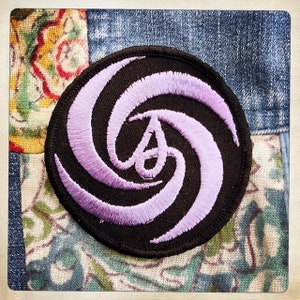 Puede incluir: Un parche bordado morado y negro con un diseño en espiral y la letra "A" en el centro.