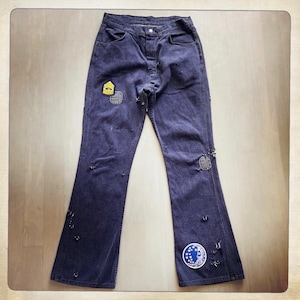 May include: Dark blue denim bell bottom pants with patches and distressed details. The patches include a yellow military insignia, a circular patch with a white star, and a blue patch with a crescent moon and stars.