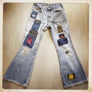 Può includere: Jeans blu a zampa d'elefante in denim con varie toppe cucite. Le toppe includono un segno di pace, un design rosso, bianco e blu, una toppa marrone, una toppa verde, una toppa blu con il testo "OLD JEANS" e una toppa gialla con il testo "LEMONS ARE YELLOW".