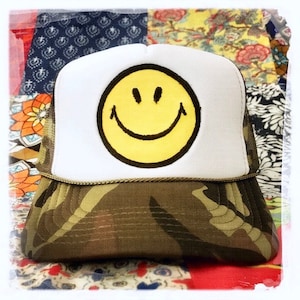 May include: A white and camouflage trucker hat with a yellow smiley face embroidered on the front.