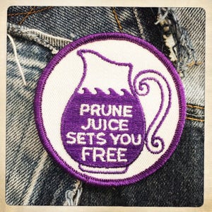 Puede incluir: Un parche bordado morado y blanco con una jarra y el texto "PRUNE JUICE SETS YOU FREE".