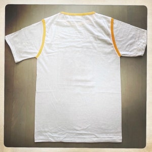 Puede incluir: Camiseta blanca con ribete amarillo alrededor del cuello y las mangas.
