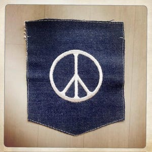Puede incluir: Un parche de tela vaquera azul oscuro con forma de bolsillo y un símbolo de la paz bordado en blanco en el centro. El parche presenta costuras blancas visibles a lo largo de los bordes y un dobladillo inferior en punta, mostrando un diseño textil clásico.