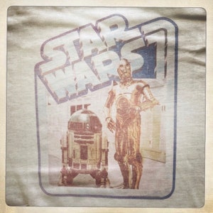 Puede incluir: Una camiseta vintage de Star Wars con una imagen descolorida de C-3PO y R2-D2. La camiseta es beige con un borde azul alrededor de la imagen. El texto "STAR WARS" est&aacute; impreso en letras grandes y en negrita en la parte superior de la imagen.