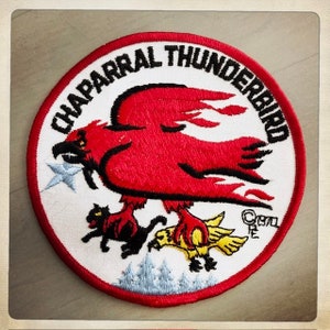 Może przedstawiać: Biała naszywka haftowana z czerwonymi, czarnymi i żółtymi detalami. Naszywka przedstawia czerwonego ptaka z czarnymi konturami i płomieniami, trzymającego żółtego ptaka w szponach. Tekst "CHPARRAL THUNDERBIRD" jest napisany w czarnym okrągłym obramowaniu wokół ptaka.