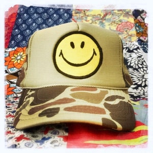 May include: A camouflage trucker hat with a yellow smiley face patch. The hat is on a colorful patchwork quilt.