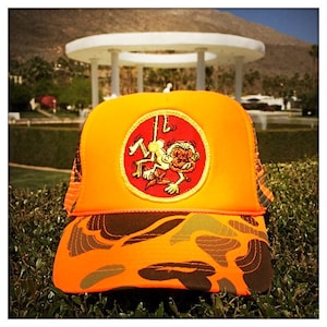 Puede incluir: Gorra de camionero de camuflaje naranja y marrón con un parche circular amarillo que presenta un mono de dibujos animados colgado de una cuerda. El mono es blanco con ojos rojos y boca roja.