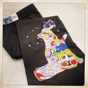 Puede incluir: Pantalones verde oscuro con un diseño bordado colorido de una mujer con un kimono japonés tradicional. El bordado presenta una variedad de colores, incluyendo rosa, amarillo, azul y verde.