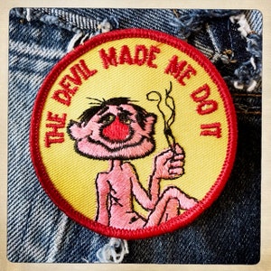 Puede incluir: Un parche bordado amarillo y rojo con un personaje de dibujos animados fumando un cigarrillo. El texto en el parche dice "The Devil Made Me Do It."