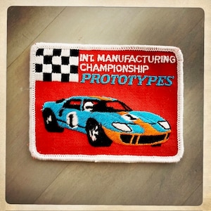 Puede incluir: Un parche bordado rojo y blanco con una bandera a cuadros, el texto "INT. MANUFACTURING CHAMPIONSHIP PROTOTYPES" y un coche de carreras azul y naranja.
