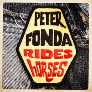 Puede incluir: Un parche hexagonal amarillo con texto bordado en negro y rojo que dice "Peter Fonda Rides Horses".