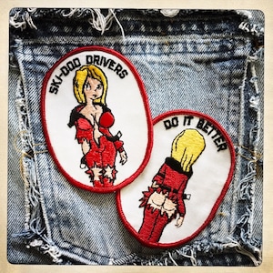 May include: Two embroidered patches on a denim background. The first patch is white with a red border and says "Ski-Doo Drivers" with a cartoon woman in a red jumpsuit. The second patch is white with a red border and says "Do It Better" with a cartoon woman in a red jumpsuit.