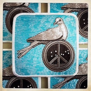 May include: A white dove with a peace sign on its back, embroidered on a blue background. The dove is perched on a brown peace sign with white dots. The patch is surrounded by a white border.