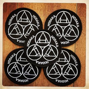 May include: Six black and white embroidered patches with the word "HODAKA" written in a circle around three overlapping triangles. The triangles are white and the background is black.