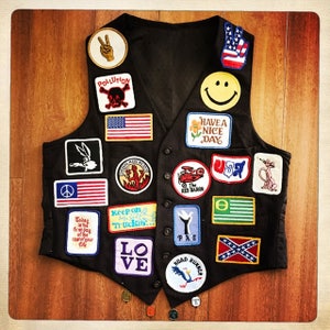 May include: A black waistcoat with a variety of patches, including peace signs, American flags, and a smiley face. The waistcoat also has patches with the words "Pollution", "Keep on Truckin'" and "Have a Nice Day".