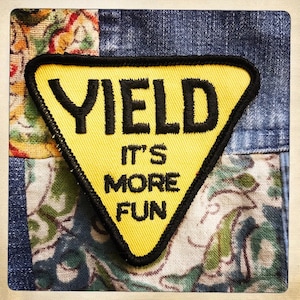 Puede incluir: Un parche triangular amarillo con costuras negras y el texto "YIELD IT'S MORE FUN".