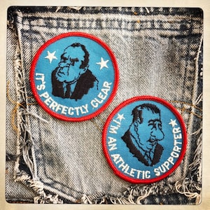 May include: Two blue and red embroidered iron-on patches with a cartoon image of a man with a large nose. The text on the patches reads "It's Perfectly Clear" and "I'm An Athletic Supporter".