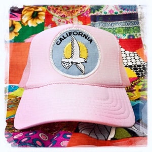 Puede incluir: Gorra de camionero rosa con un parche azul y blanco que dice "CALIFORNIA" con una paloma blanca volando frente a un sol amarillo.