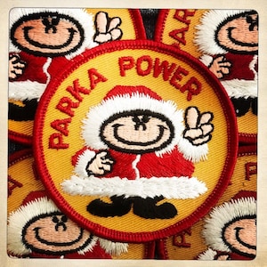 May include: A yellow and red embroidered patch featuring a cartoon character in a red and white Santa suit, making a peace sign with two fingers. The text "Parka Power" is written around the figure.