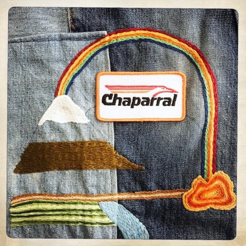 Puede incluir: Una chaqueta vaquera con detalles bordados, incluyendo un arco&iacute;ris, una monta&ntilde;a, una nube y un parche que dice "Chaparral".