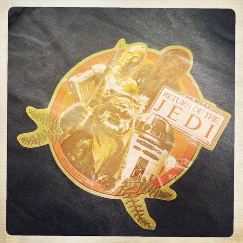Op de afbeelding: Een vintage Star Wars T-shirt met een cirkelvormig ontwerp met personages uit de film Return of the Jedi. De personages zijn Chewbacca, R2-D2 en C-3PO. Het ontwerp is in tinten geel, oranje en bruin op een zwarte achtergrond.