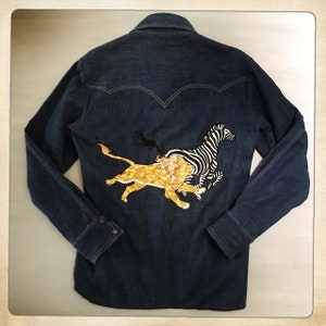 May include: A dark blue denim shirt with an embroidered design of a lion chasing a zebra on the back.